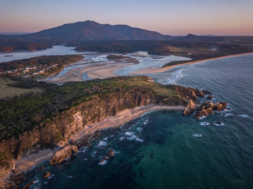 Aerial view of a rocky headland and curving beach, with Gulaga rising behind