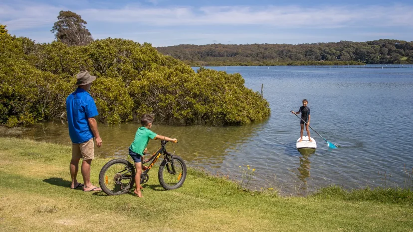 A family with bikes on the riverbank, watching a stand-up paddleboarder