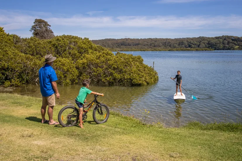 A family with bikes on the riverbank, watching a stand-up paddleboarder