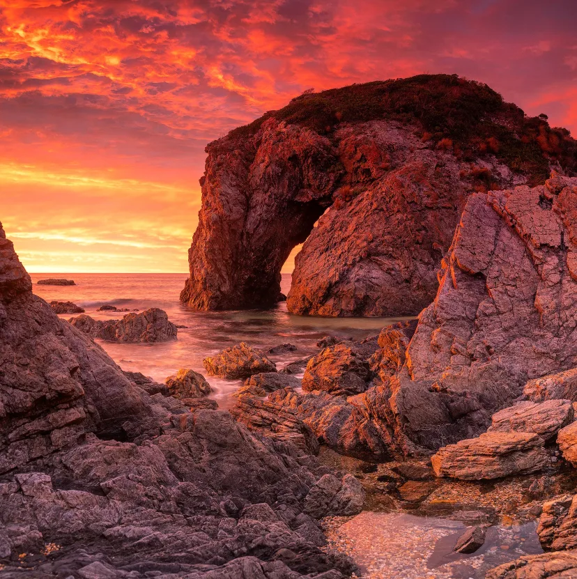 Horse Head Rock at sunset, the sea arch silhouetted against a deep red sky with the tide washing through the rock platform