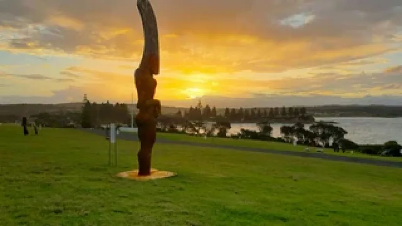 A tall dark sculpture on the Bermagui headland at sunset, with the harbour behind