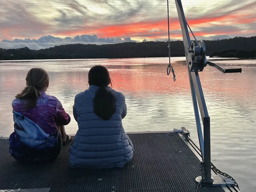 Two people sitting on the jetty at sunset beside a moored boat